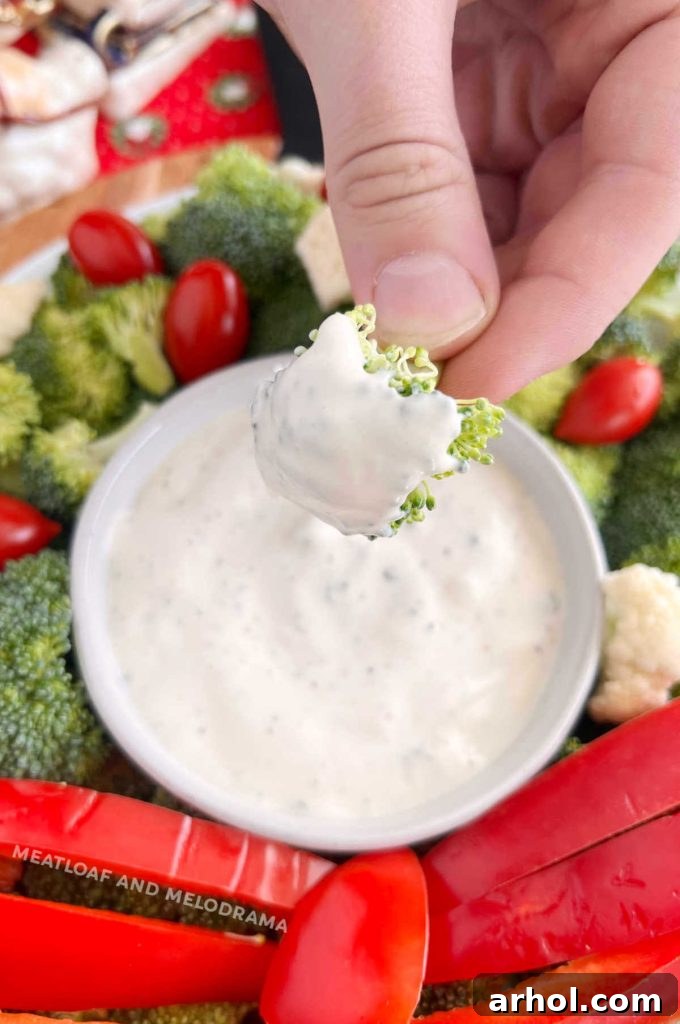 Close-up of a hand holding a piece of fresh broccoli, perfectly dipped in ranch dressing, against a blurred festive background.