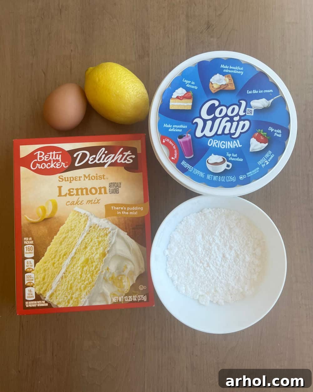 A flat lay of all the essential ingredients for lemon cake mix crinkle cookies: a yellow box of lemon cake mix, a fresh lemon, a large egg, a container of Cool Whip, and a bowl of powdered sugar, all arranged on a rustic wooden board.