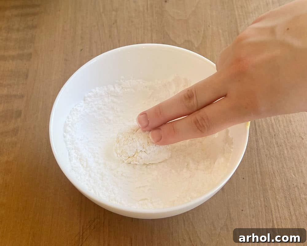 A hand gently rolling a sticky ball of lemon cookie dough in a bowl of powdered sugar until it is fully coated, ready for baking.