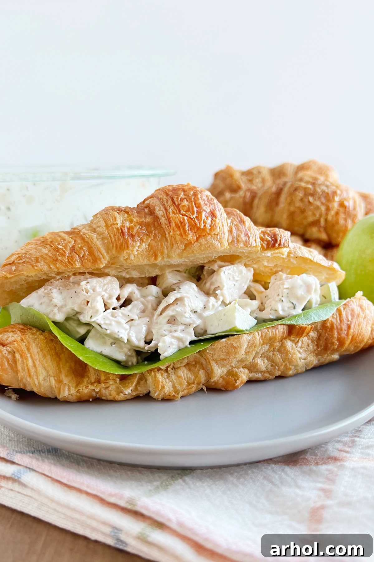 Fresh Lemon Chicken Salad with Apples served in a flaky croissant roll on a pristine white plate, garnished with a sprinkle of fresh herbs.