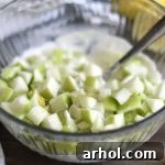 A bowl containing a creamy chicken salad dressing, with freshly diced green apples being stirred in, highlighting the initial steps of combining ingredients.