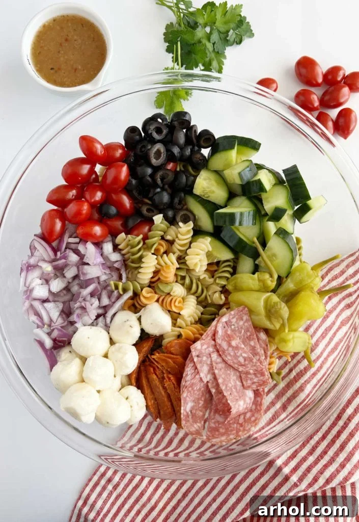 All the fresh ingredients for pepperoni pasta salad recipe laid out in separate bowls: rotini pasta, cucumber, grape tomatoes, mozzarella balls, black olives, pepperoncini, red onion, salami, and pepperoni.