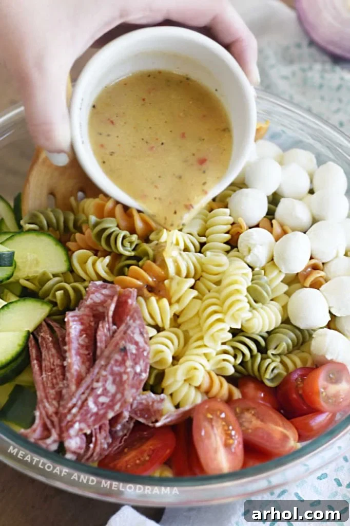 Zesty Italian dressing being poured over the colorful pasta, pepperoni, salami, and vegetables in a large mixing bowl.