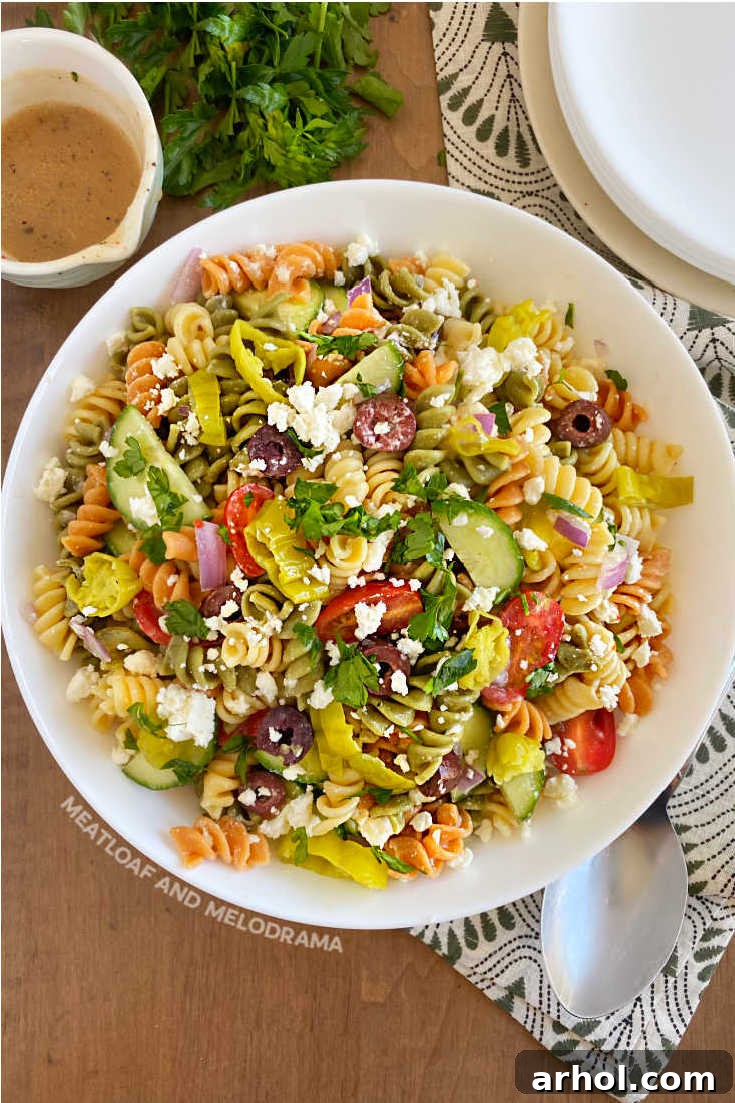 Overhead shot of vibrant Greek pasta salad with tri-color rotini, feta, olives, and fresh vegetables in a white serving bowl, ready for a summer gathering.