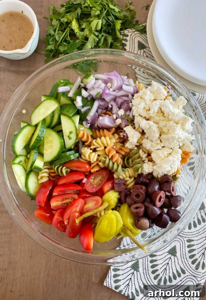 Assortment of fresh Greek pasta salad ingredients: rotini, Kalamata olives, crumbled feta, sliced cucumbers, grape tomatoes, red onion, and pepperoncini.