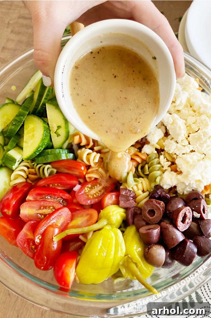 Pouring zesty homemade dressing over a bowl of tri-color pasta, fresh vegetables, and feta cheese for a Greek pasta salad.