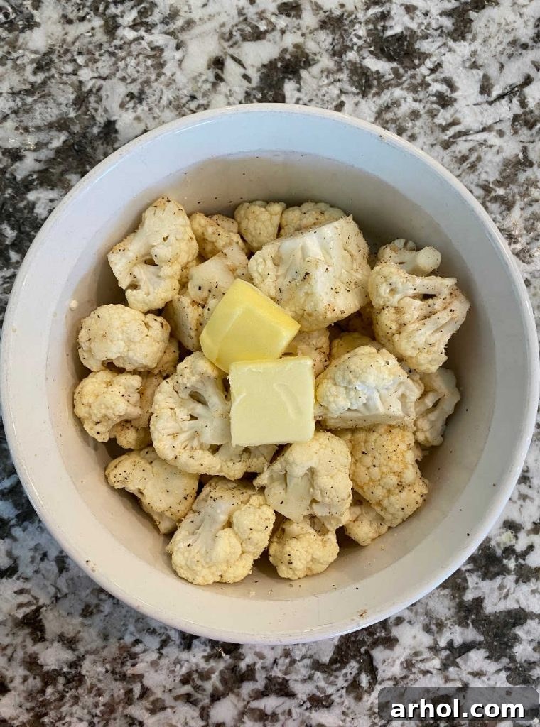 Raw cauliflower florets seasoned with salt, pepper, garlic powder, and butter in a microwave-safe dish.