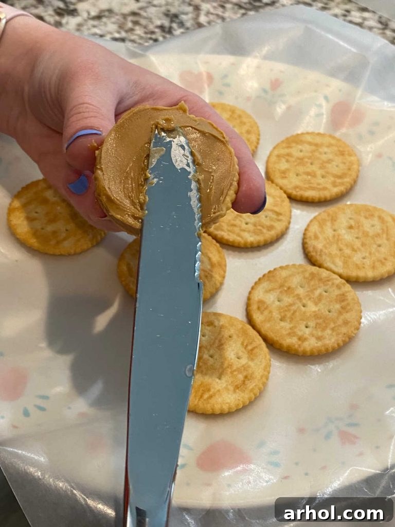 A hand using a knife to spread creamy peanut butter meticulously onto a round Ritz cracker, preparing the base for the sandwich cookie.