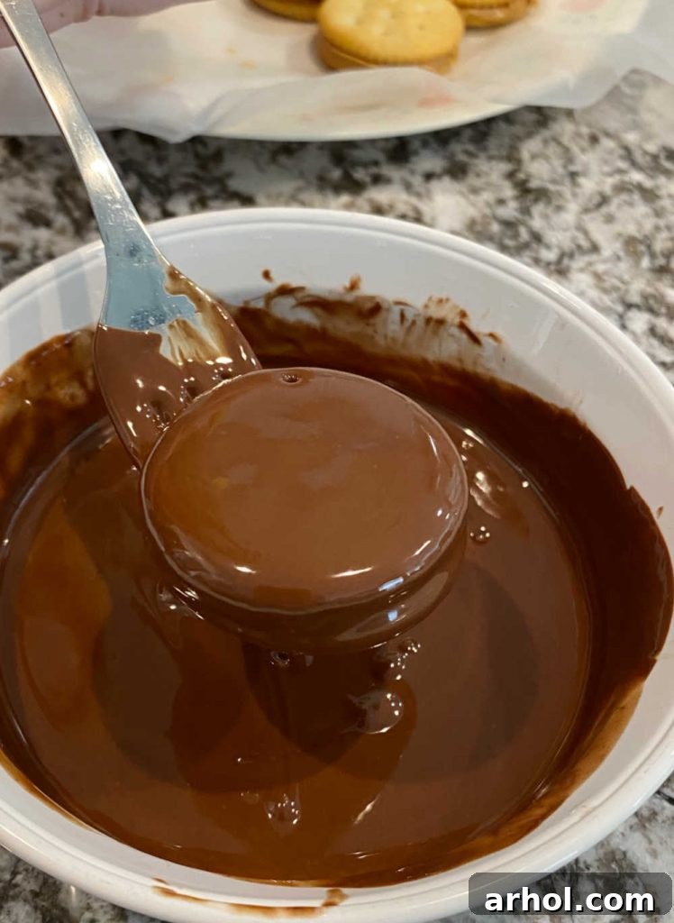 A Ritz peanut butter cracker sandwich being carefully dipped into a bowl of perfectly melted chocolate using a fork, ensuring full coverage.