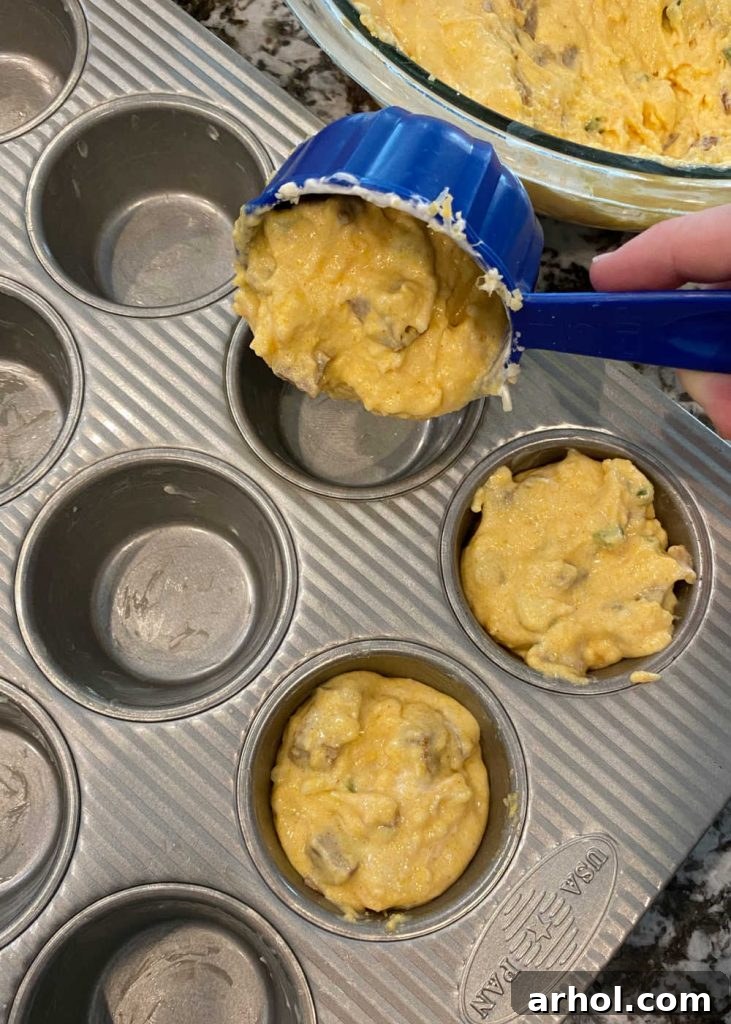 Cornbread muffin batter being carefully spooned into a prepared muffin tin, with some cups already topped with a decorative jalapeño slice.