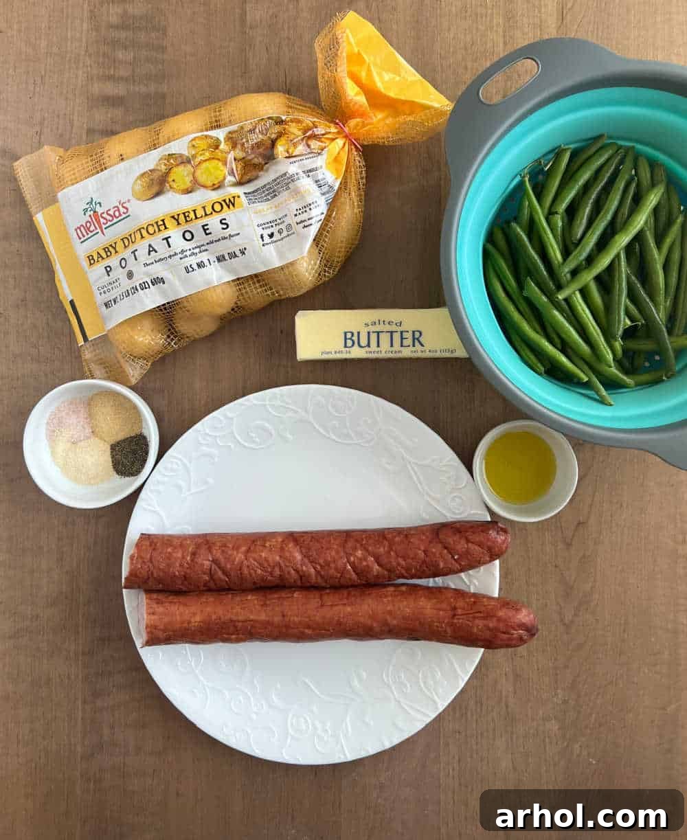 Ingredients for slow cooker sausage and potatoes: smoked sausage, baby dutch potatoes, fresh green beans, butter, olive oil, and various spices laid out on a surface.