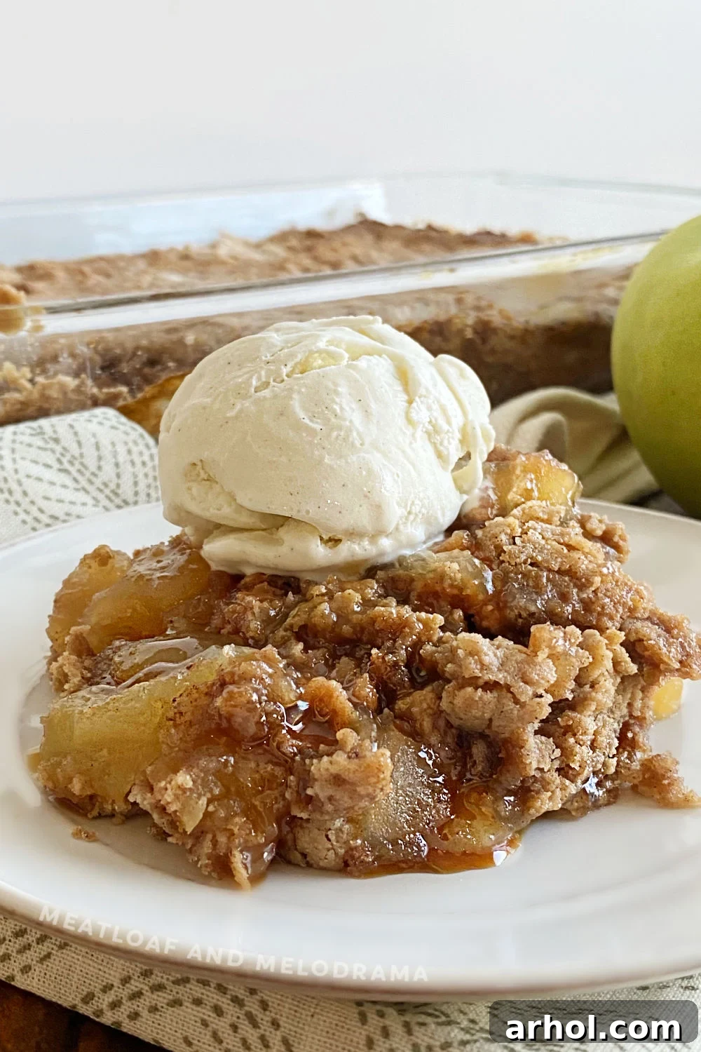 Warm apple pie dump cake with a scoop of melting vanilla ice cream and caramel drizzle on a white plate, garnished with a thin apple slice.