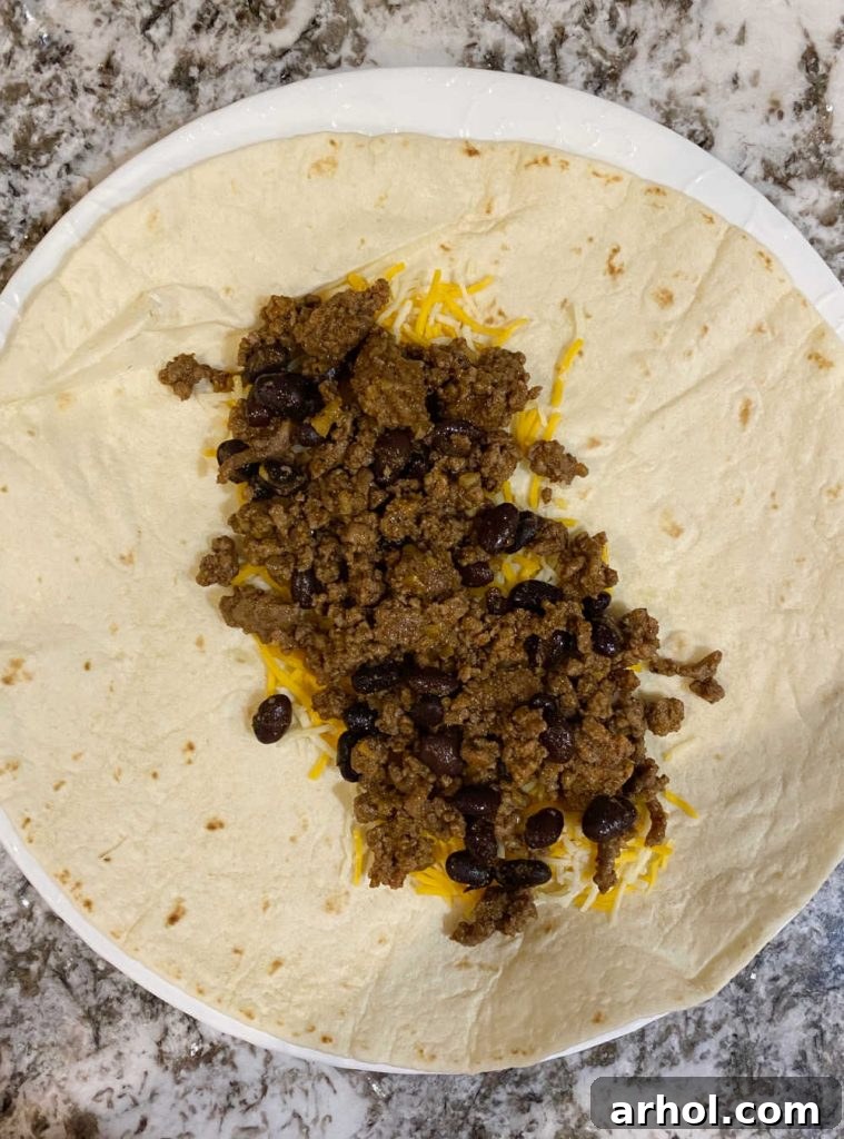 A large flour tortilla with ground beef and black bean filling, topped with shredded cheddar cheese, prepared for rolling into a wet burrito.