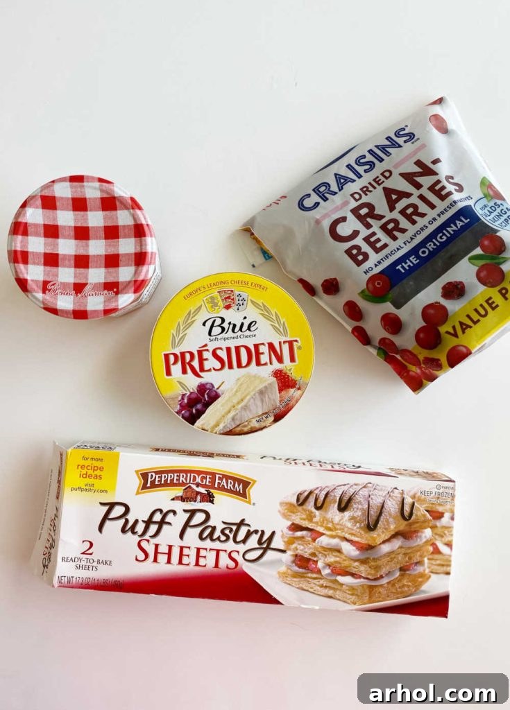 Ingredients for baked brie: puff pastry sheet, wheel of brie cheese, orange marmalade, and dried cranberries displayed on a wooden table.