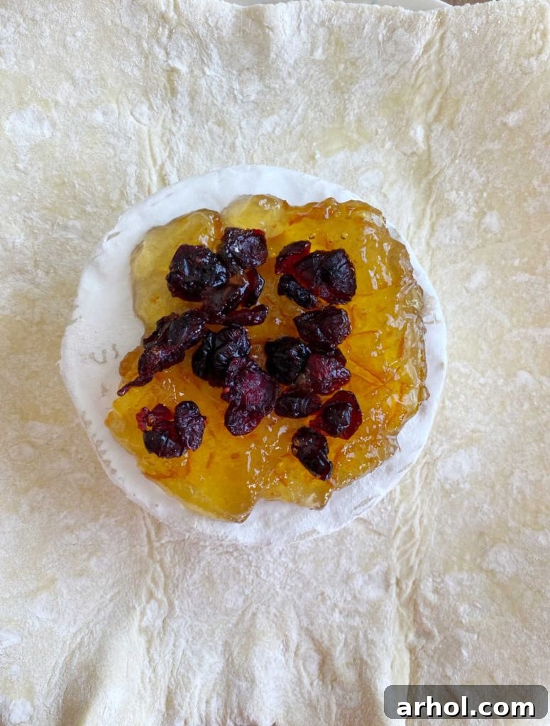A wheel of brie cheese placed on a puff pastry sheet, topped with vibrant orange marmalade and dried cranberries, ready for wrapping.