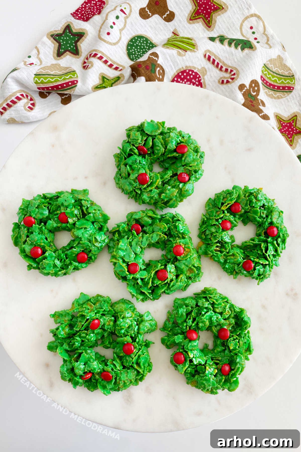 Festive Christmas cornflake wreath cookies adorned with red M&M candies, elegantly displayed on a marble cake stand, capturing the spirit of holiday baking and easy no-bake treats.