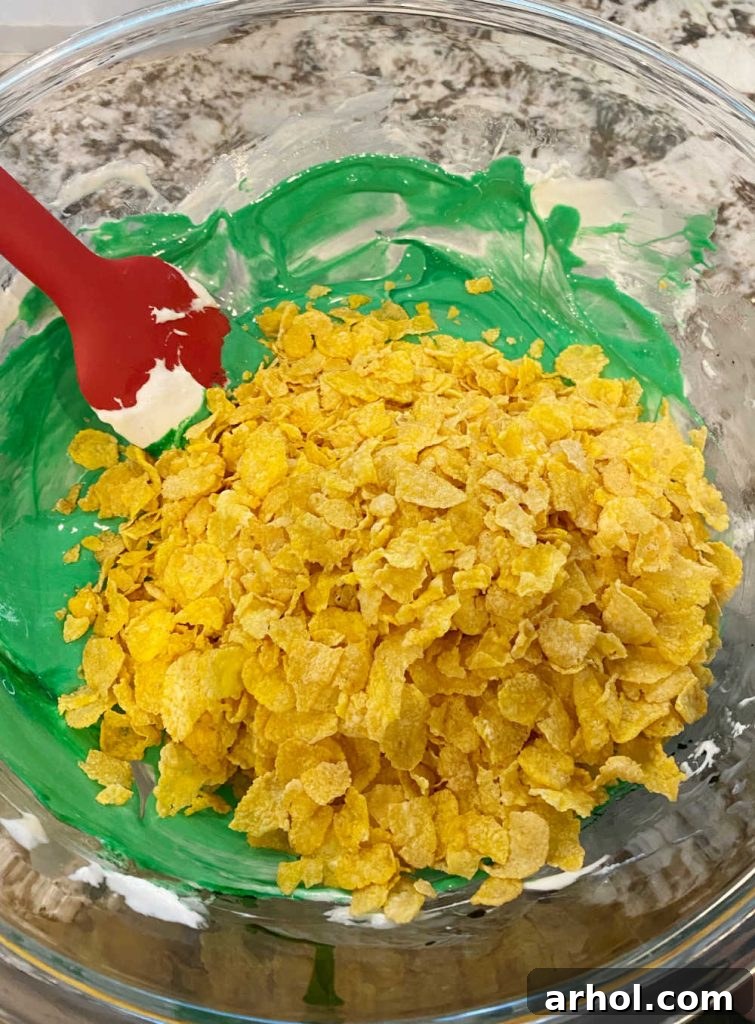 A close-up of a large mixing bowl where crisp cornflakes are being stirred into a vibrant green marshmallow mixture, illustrating the gentle folding required for Christmas cornflake wreath cookies.