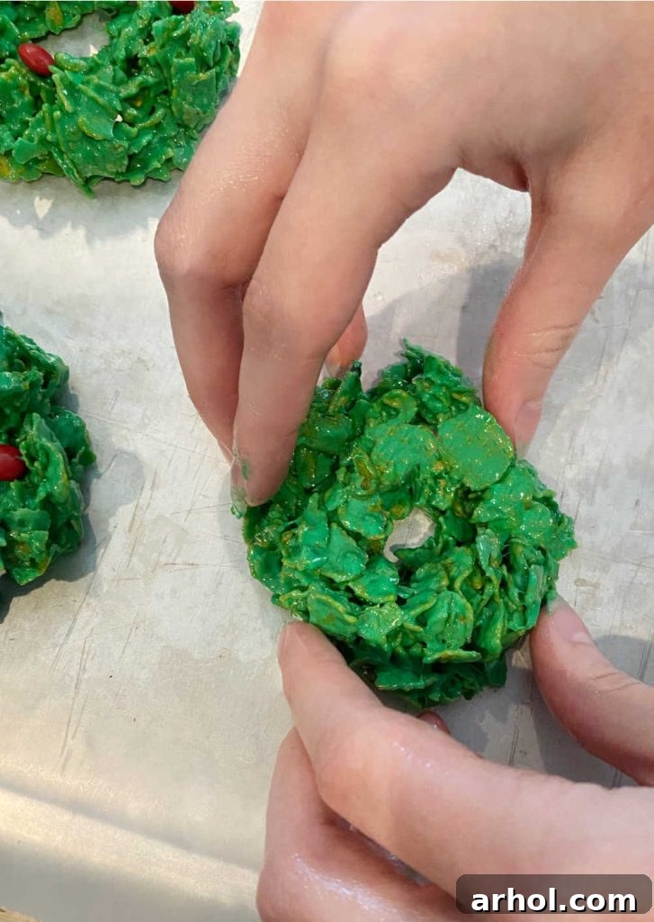 Hands delicately forming a green cornflake mixture into small wreath shapes on a sheet of parchment paper, highlighting the hands-on process of making Christmas cornflake wreath cookies.