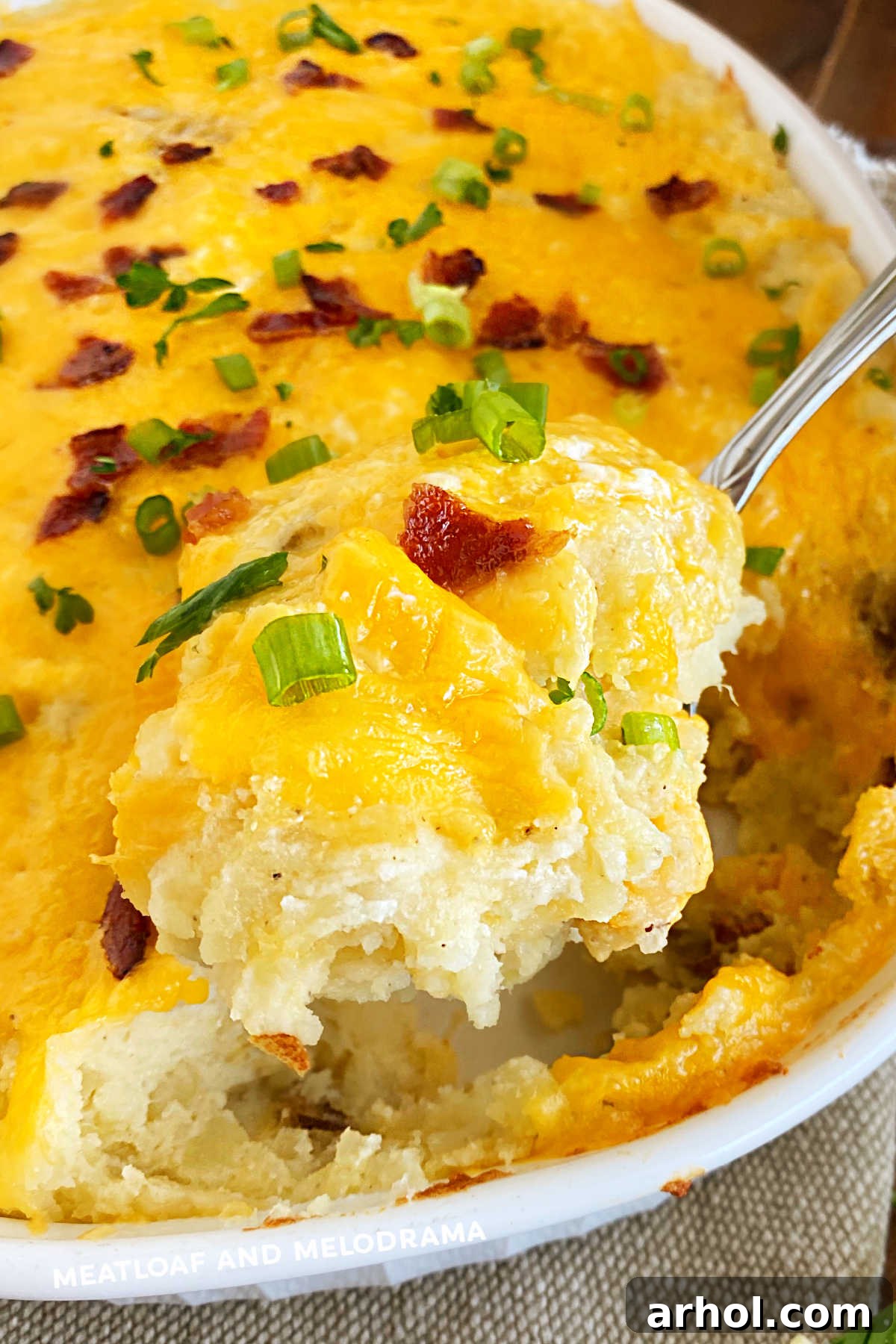 A serving spoon lifting a portion of twice baked potatoes casserole topped with golden cheddar cheese and crispy bacon, ready to be enjoyed.