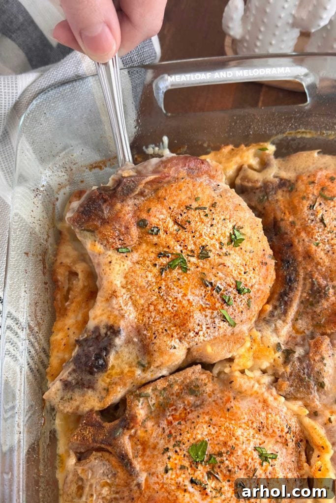 A hand lifting a perfectly baked pork chop out of a baking dish filled with creamy rice, showing the tenderness and succulence of the cooked meal.