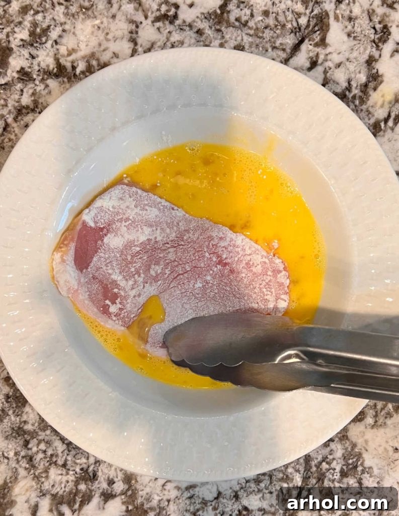 A pork cutlet, previously coated in flour, being dipped into beaten eggs using kitchen tongs, showing the three-step breading process.