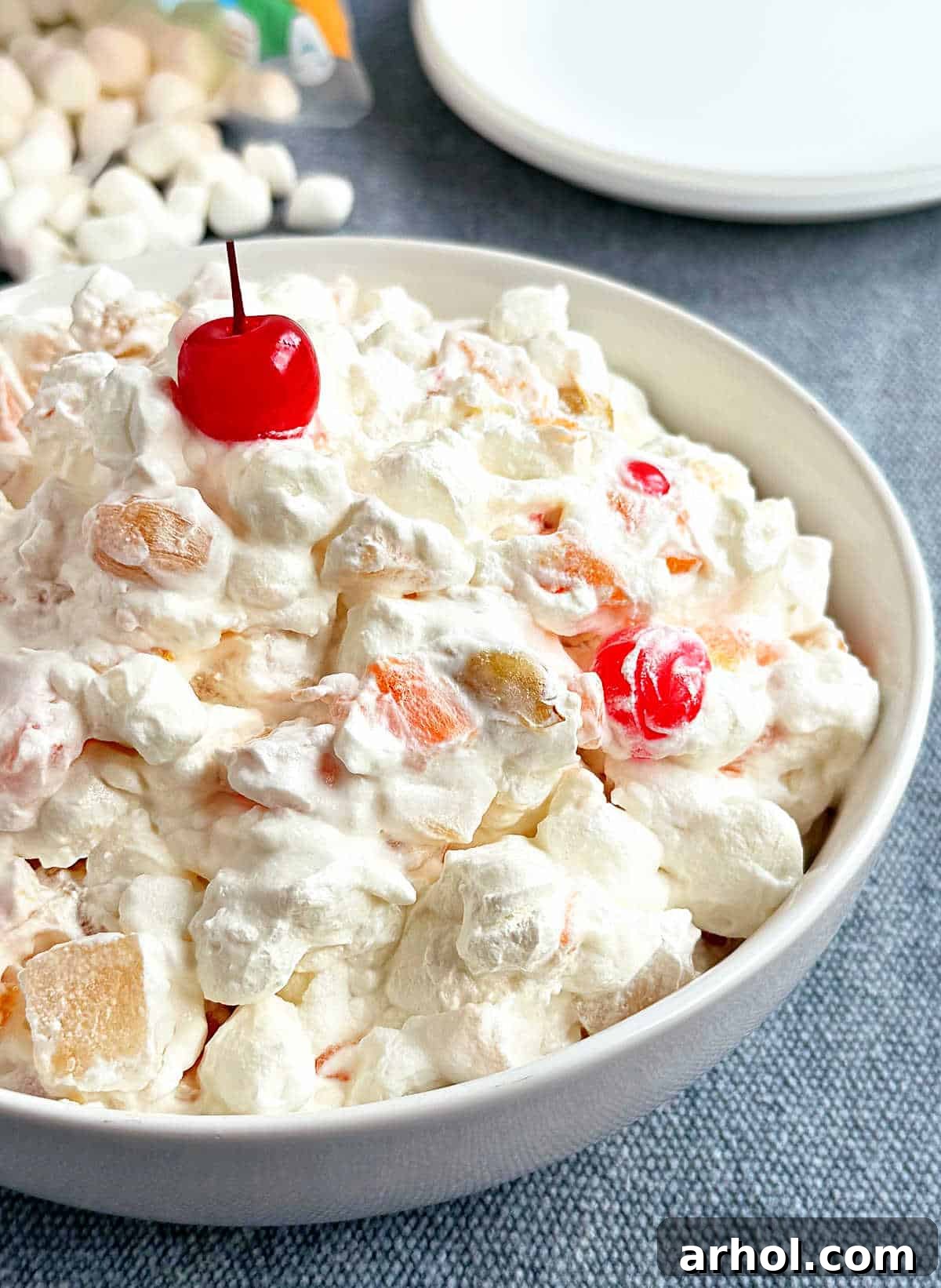 A large bowl of Marshmallow Fruit Salad with a vibrant red maraschino cherry on top, sitting on a rustic wooden surface, ready to be served.