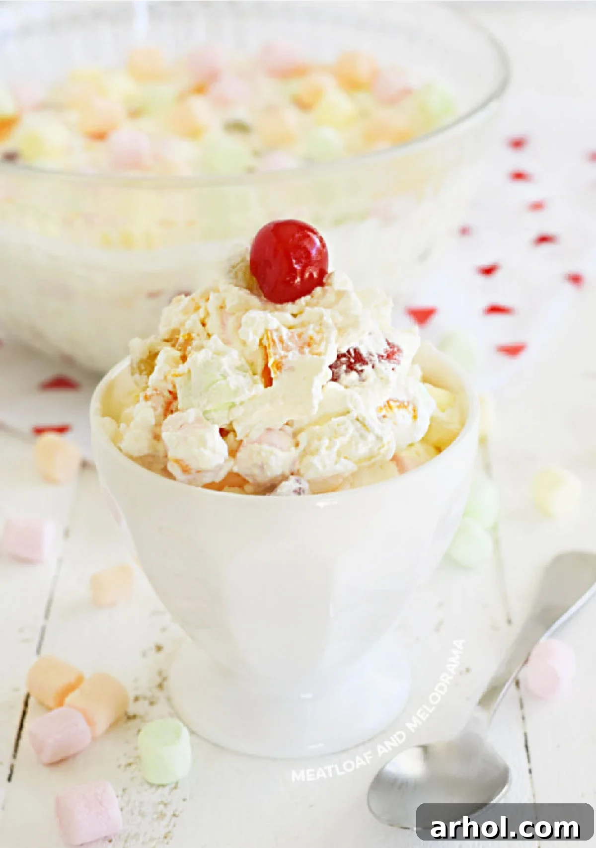 A white serving dish filled with creamy Marshmallow Fruit Salad, garnished with a single maraschino cherry and placed on a light-colored wooden table.