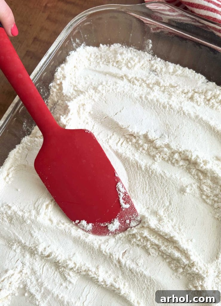 Dry yellow cake mix being smoothed over the strawberry pie filling in a baking pan using a red spatula.