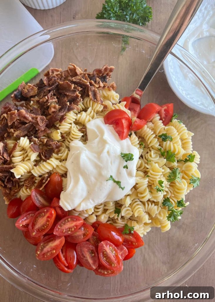 Perfect BLT Pasta Salad 5 Creamy mayonnaise ranch dressing being mixed with rotini pasta, crispy bacon, and juicy tomatoes in a large glass bowl.