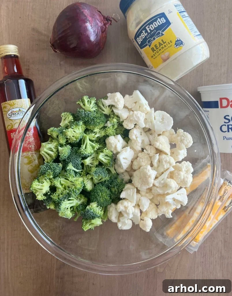 An array of fresh ingredients for broccoli cauliflower salad: vibrant broccoli florets, white cauliflower florets, creamy mayonnaise, tangy sour cream, sharp red onion, cubed cheese, and a bottle of red wine vinegar