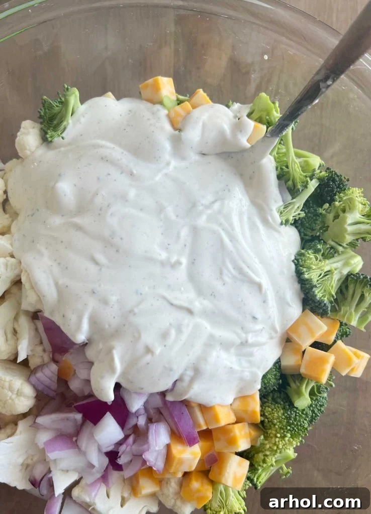 The creamy mayonnaise and sour cream dressing being poured over the colorful broccoli, cauliflower, red onion, and cheese mixture in a large mixing bowl