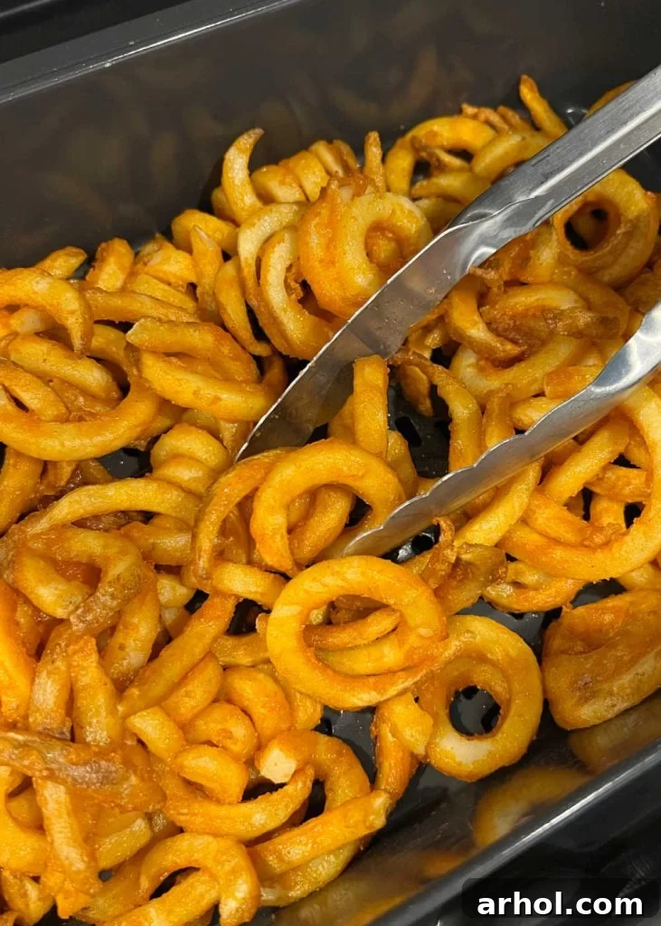 A person using kitchen tongs to turn and shake frozen curly fries in an air fryer basket during cooking for even crisping.
