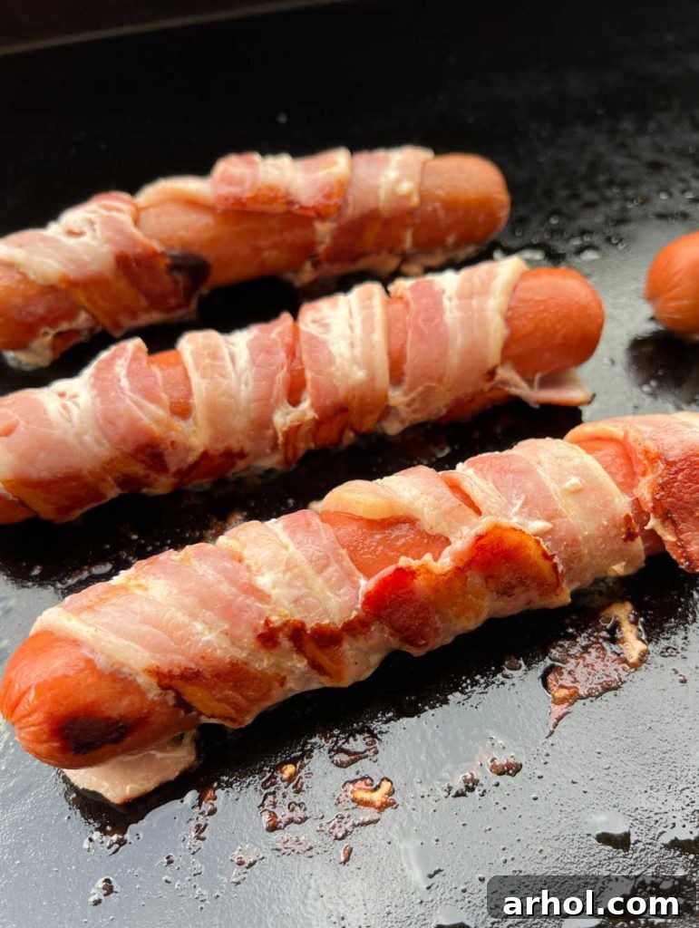 Authentic Sonoran Hot Dogs Blackstone Griddle Style 5 Bacon-wrapped hot dogs sizzling and cooking on a hot Blackstone griddle, achieving a perfect golden crisp.