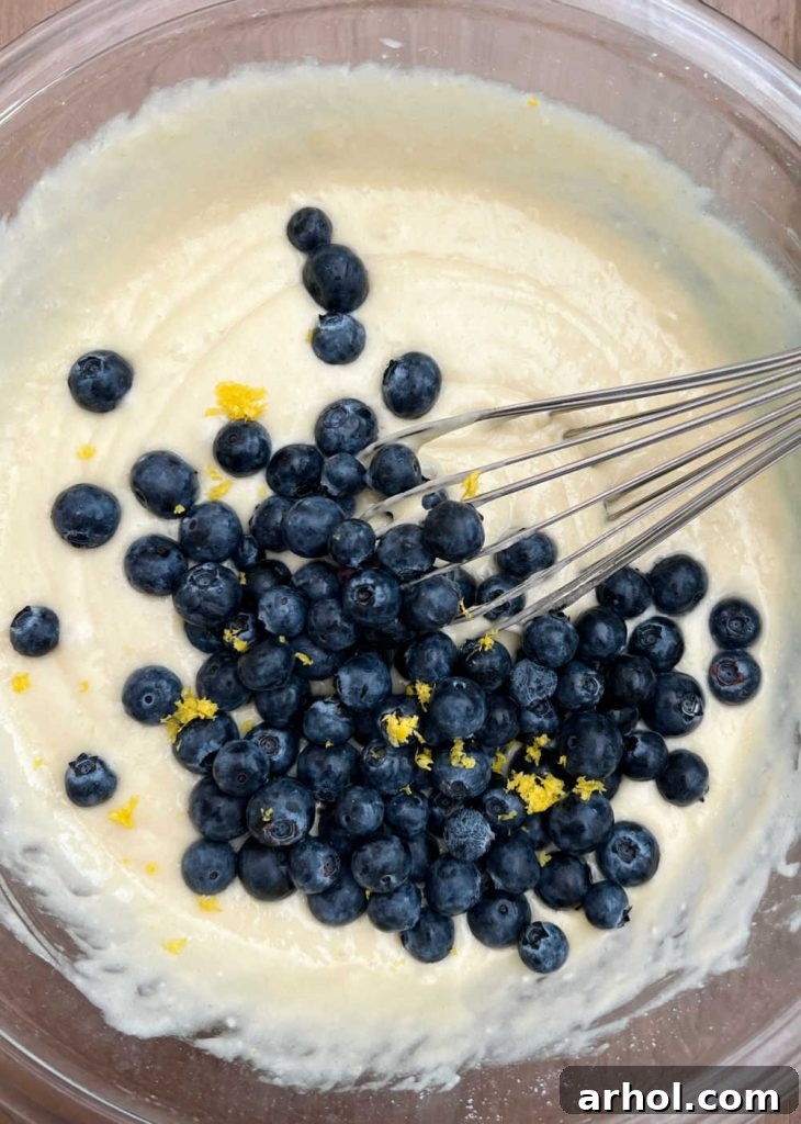 Incorporating Blueberries and Lemon Zest Fresh blueberries and lemon zest being gently folded into the thick muffin batter with a rubber spatula