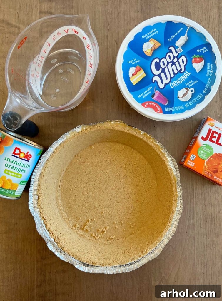 Key ingredients for orange creamsicle pie laid out: a box of orange Jello, a tub of Cool Whip, a pitcher of water, a ready-made graham cracker pie crust, and a bowl of fresh mandarin oranges.