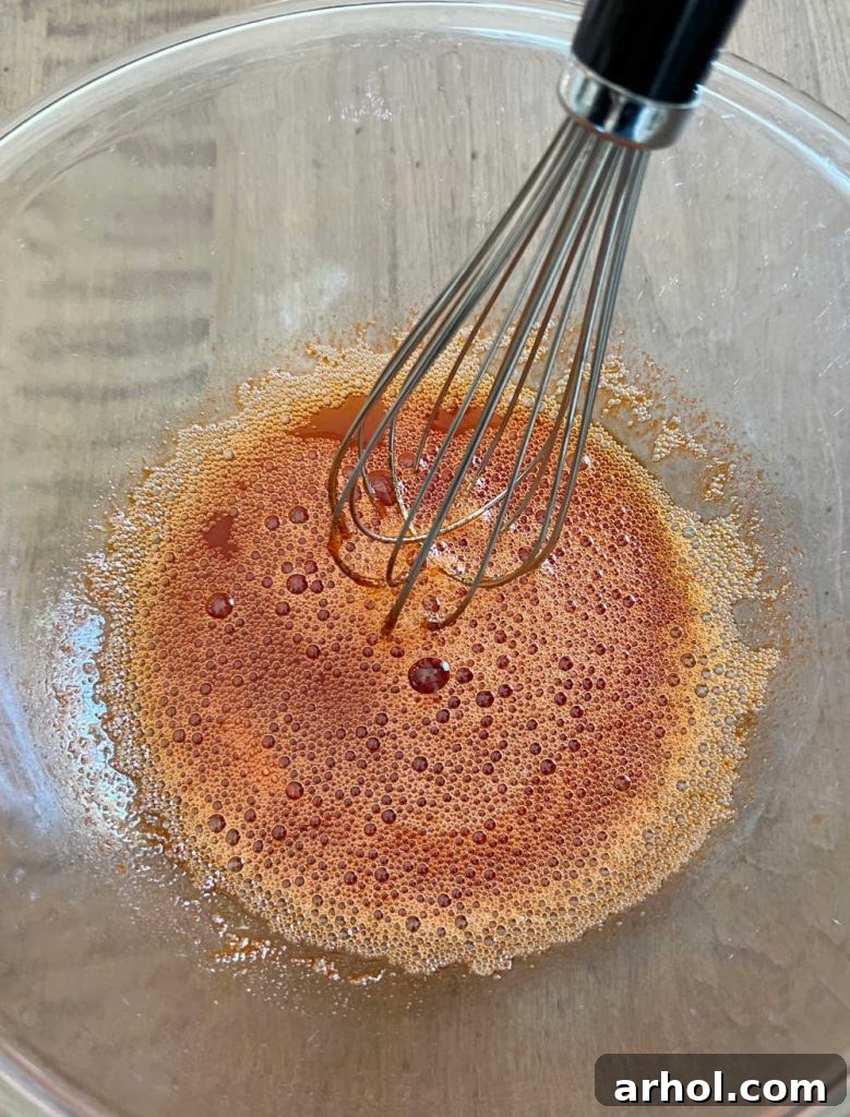 A hand whisking orange Jello gelatin powder with boiling water in a clear glass mixing bowl until fully dissolved.