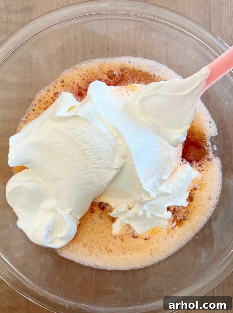A spoon gently mixing creamy Cool Whip into the pale orange Jello mixture in a glass bowl, creating a smooth, blended filling.