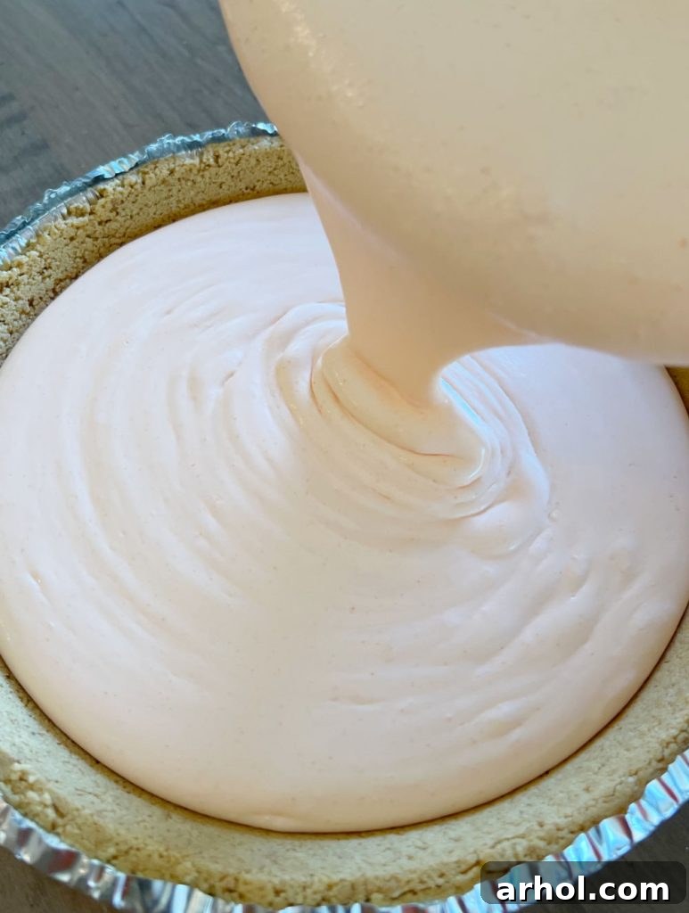 A rich, pale orange creamsicle filling being carefully spooned into a golden brown graham cracker pie crust.
