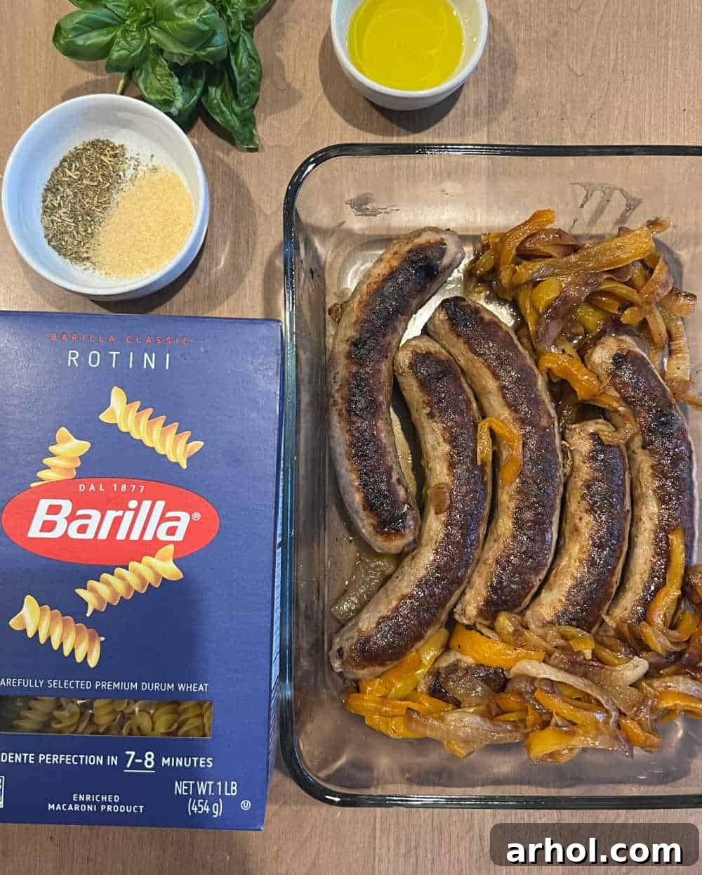 Grilled Italian sausages, colorful bell peppers and onions, pasta, fresh basil, olive oil, and seasoning laid out for a recipe.