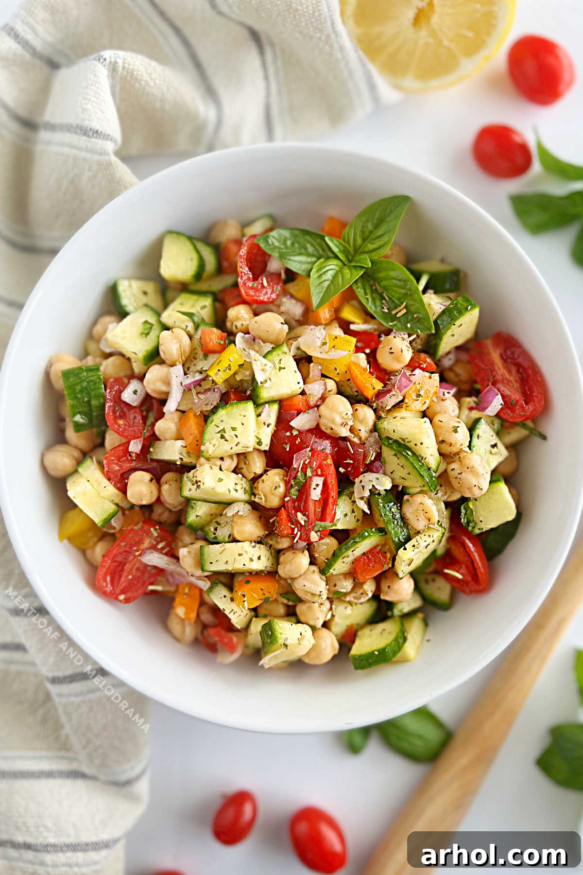 A simple and vibrant chickpea salad in a white bowl, garnished with fresh herbs, featuring chopped cucumbers, red onion, colorful bell peppers, and grape tomatoes.