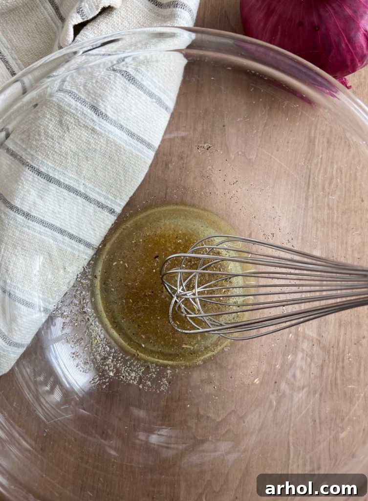 A hand whisking together lemon juice, red wine vinegar, and olive oil in a clear glass mixing bowl to create the zesty dressing for the chickpea salad.