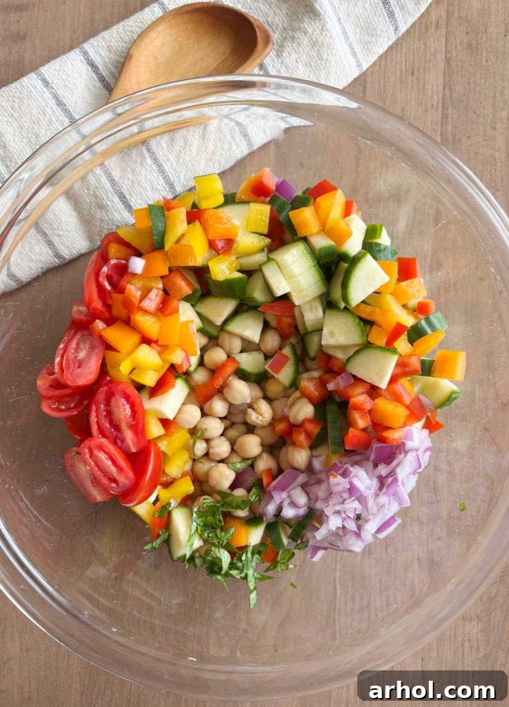 A large mixing bowl filled with vibrant, freshly chopped vegetables including cucumbers, bell peppers, grape tomatoes, red onion, and chickpeas, ready to be tossed with dressing.