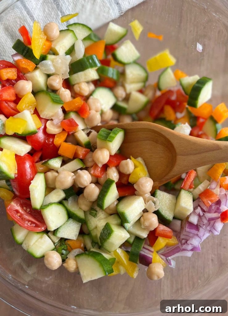 A wooden spoon mixing garbanzo beans and other colorful salad ingredients in a large bowl, ensuring everything is thoroughly combined with the dressing.