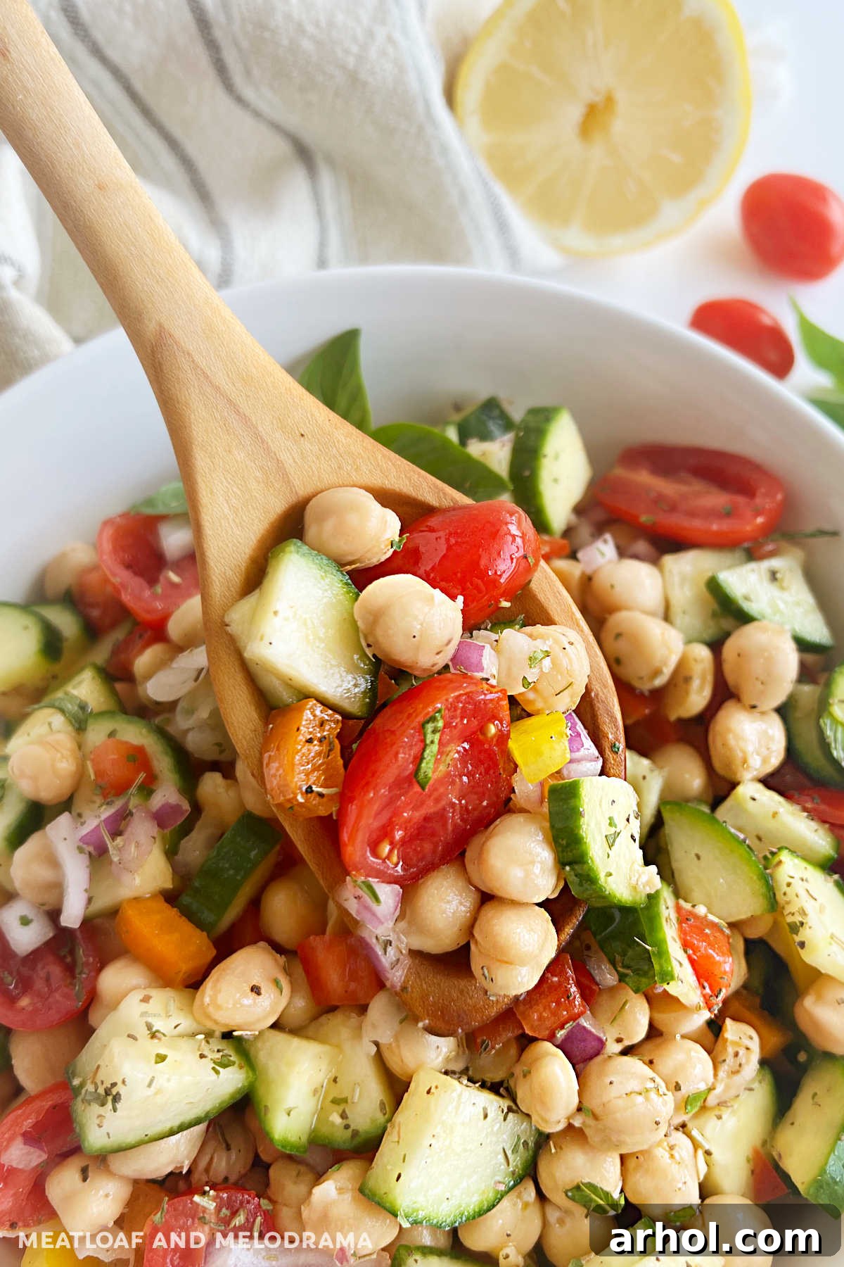 A spoonful of vibrant chickpea salad held over a white serving bowl, showcasing the colorful medley of garbanzo beans, cucumbers, tomatoes, and bell peppers.