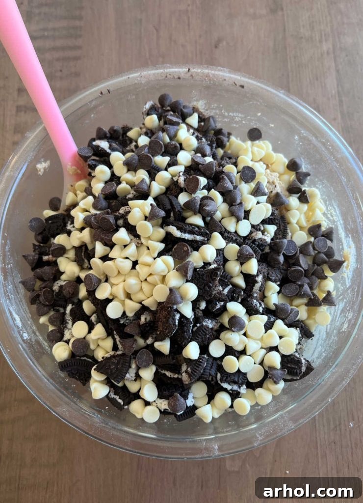 Stirring in chopped Oreo cookie pieces and various chocolate chips into the cookie dough with a spatula.