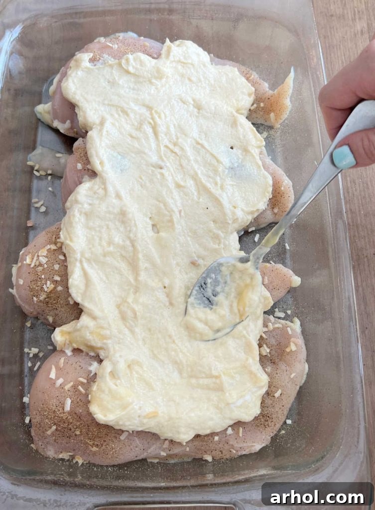 Cream of chicken soup and sour cream mixture being smoothly spread over seasoned chicken in a casserole dish, forming a creamy layer.