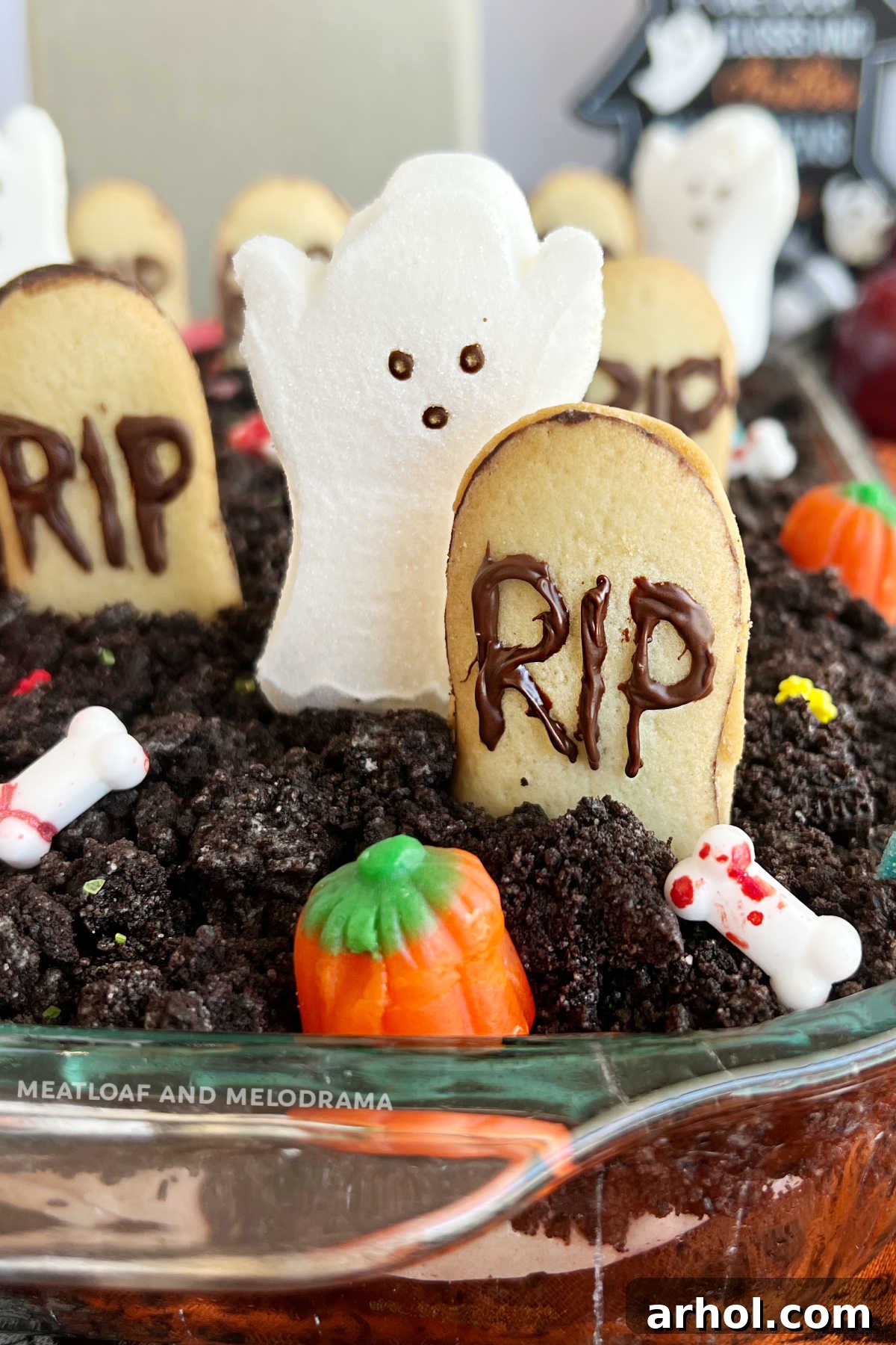 A beautifully decorated Halloween Dirt Cake (graveyard cake) in a baking dish, featuring marshmallow ghosts, candy pumpkins, gummy worms, and cookie gravestones, set against a dark, moody background perfect for Halloween.