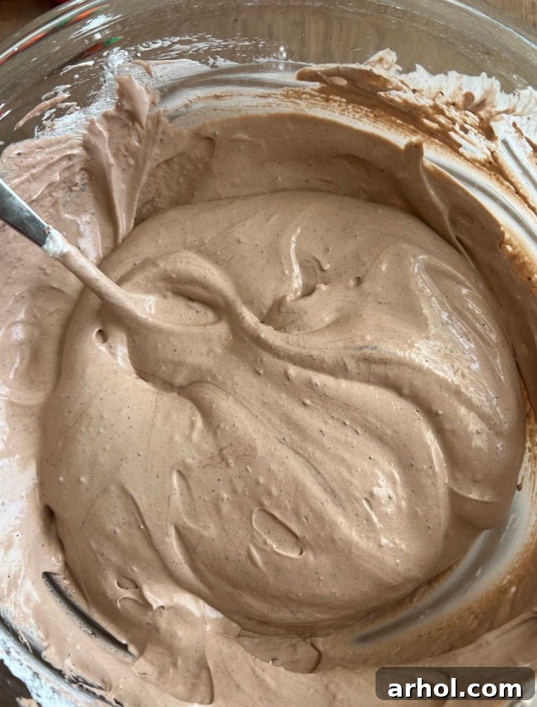 A close-up image showing a creamy, light chocolate pudding and whipped topping mixture being prepared in a large bowl, ready to be spread over the cake base.