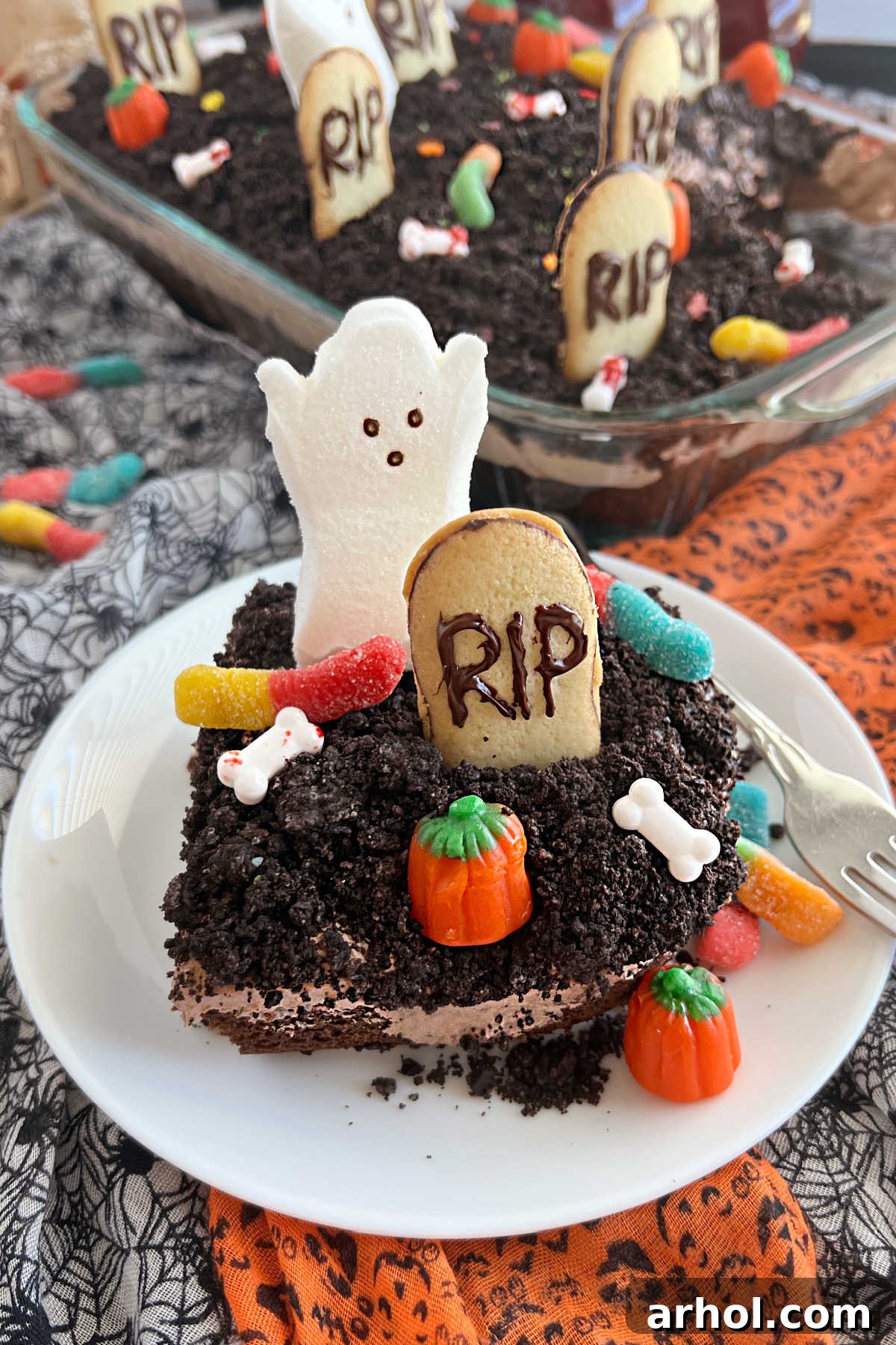A slice of Halloween Dirt Cake served on a white plate, showcasing the distinct layers of chocolate cake, pudding, and Oreo crumbs, adorned with gummy worms, cookie gravestones, and marshmallow ghost Peeps, ready to be enjoyed.