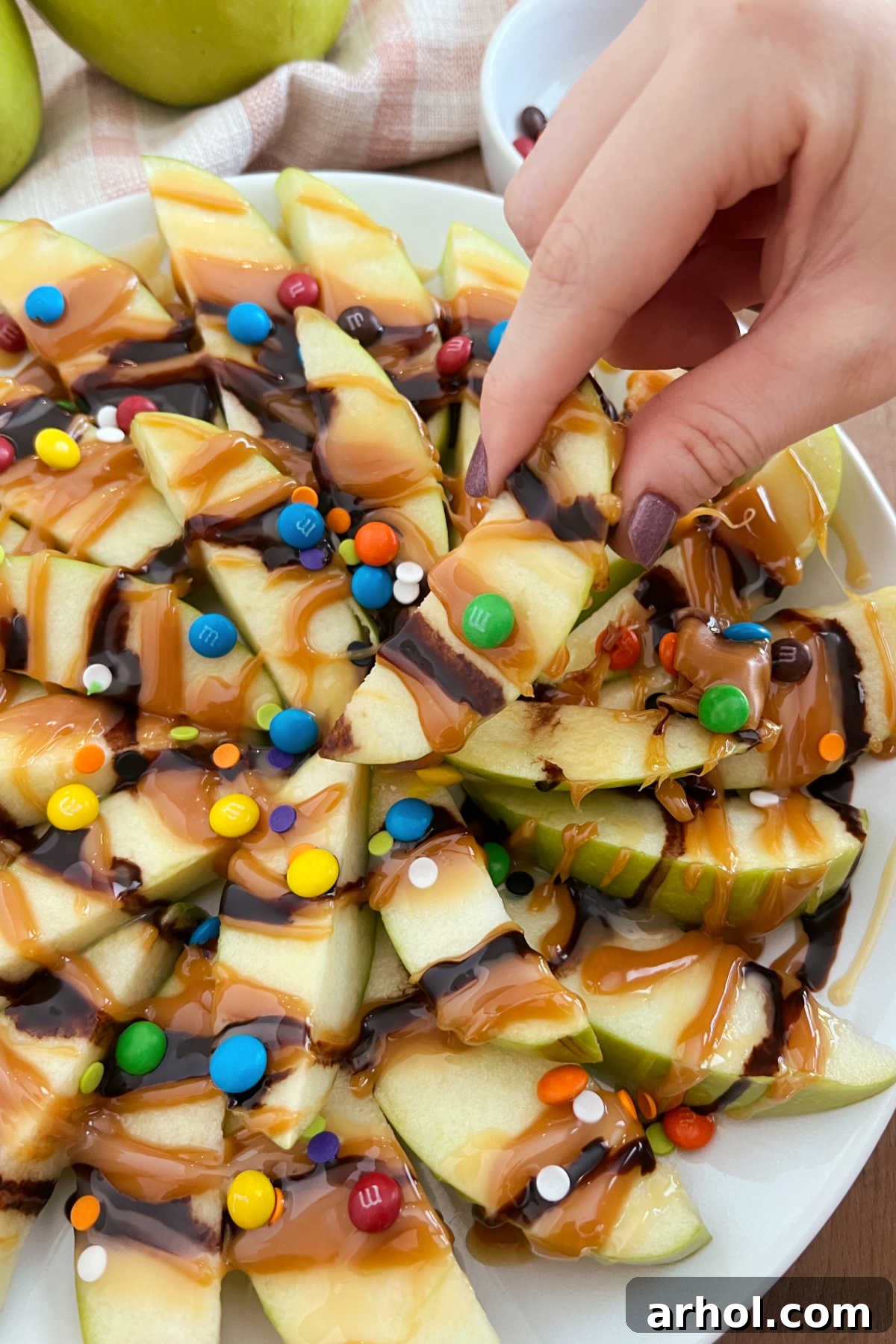 Caramel Apple Nacho Delight 6 A hand gently reaching for a perfectly prepared slice of caramel apple nacho from a full platter, highlighting the delectable layers of apple, caramel, and assorted toppings, ready for enjoyment.